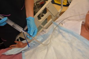 A nursing student demonstrates flushing an enteral tube with water using a practice mannequin. The student wears blue nitrile gloves while carefully pushing water from a syringe into the end of an enteral feeding tube.