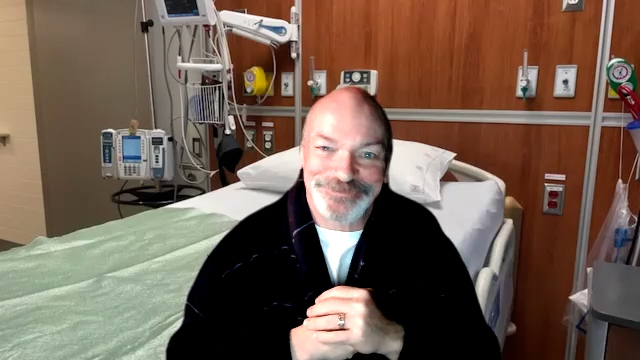 Carson Deluca, your patient. He is an adult man with greying facial hair and a shaved head. His hands are folded as he sits near his bed in the hospital, smiling warmly. He wears a black sweater over a white shirt.