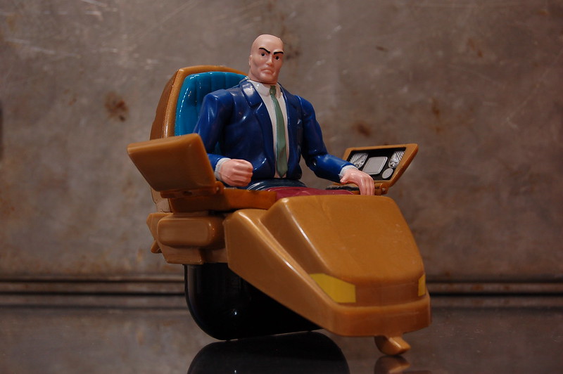 An action figure of Professor X from the X-Men by Marvel Comics. A bald white man sits in a futuristic hovering orange wheelchair. There are two pads attached to the wheelchair with screens and various interfaces near his hands.