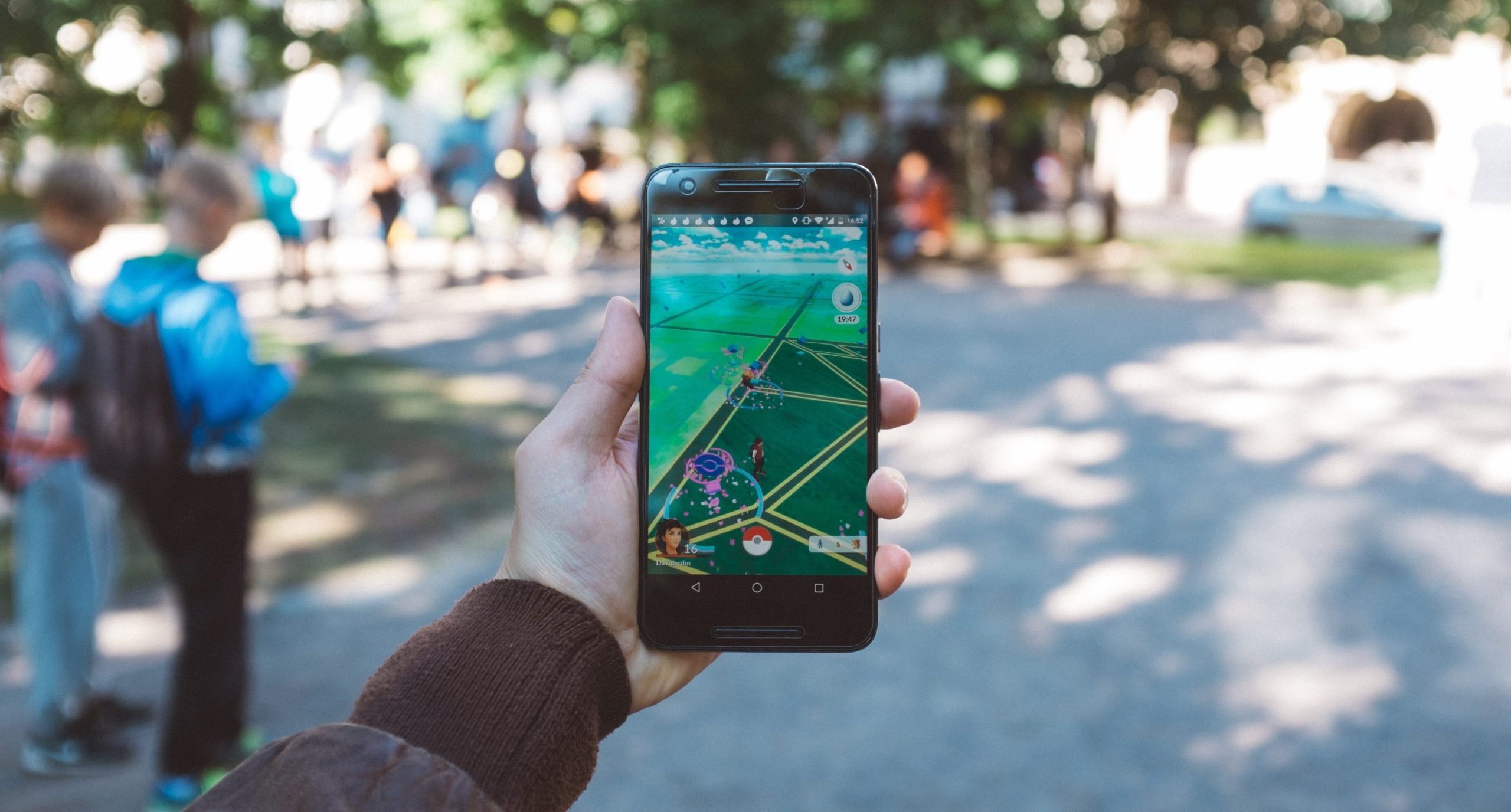 A hand holding a cell phone displaying the Pokemon GO app on map view. They are outside in a place with trees, cement and dappled sunlight.
