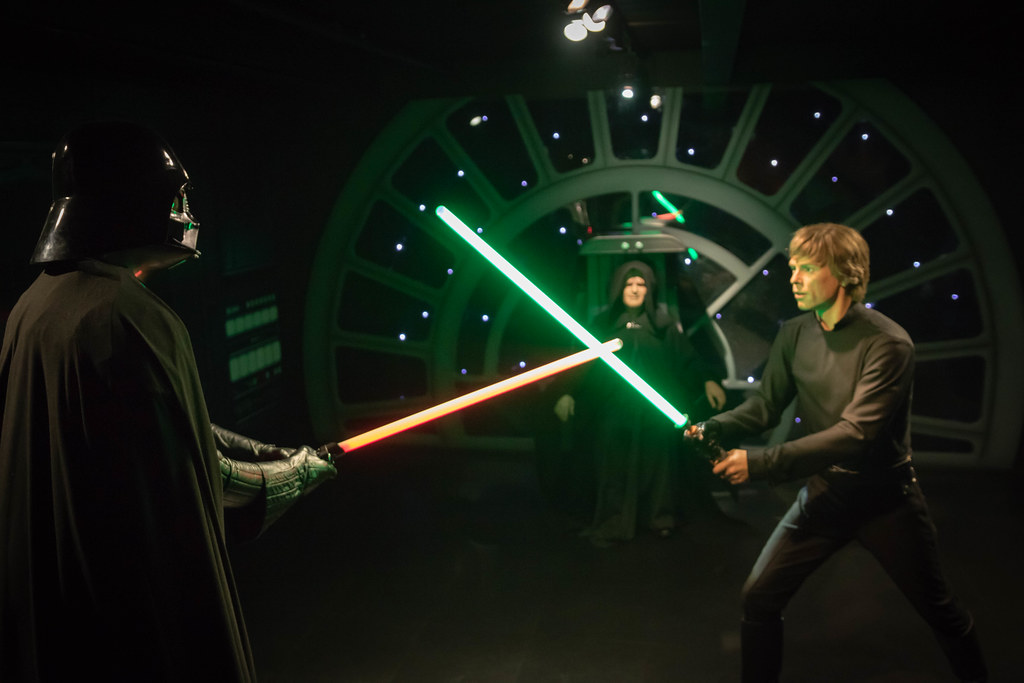 A screenshot from Star Wars where Darth Vader and Luke Skywalker battle with lightsabers in front of the Emperor Darth Sidious.