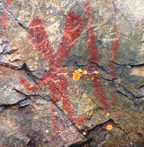 An image of what appears to be a person with two elongated heads holding a stick painted with red paint on the surface of a rock.